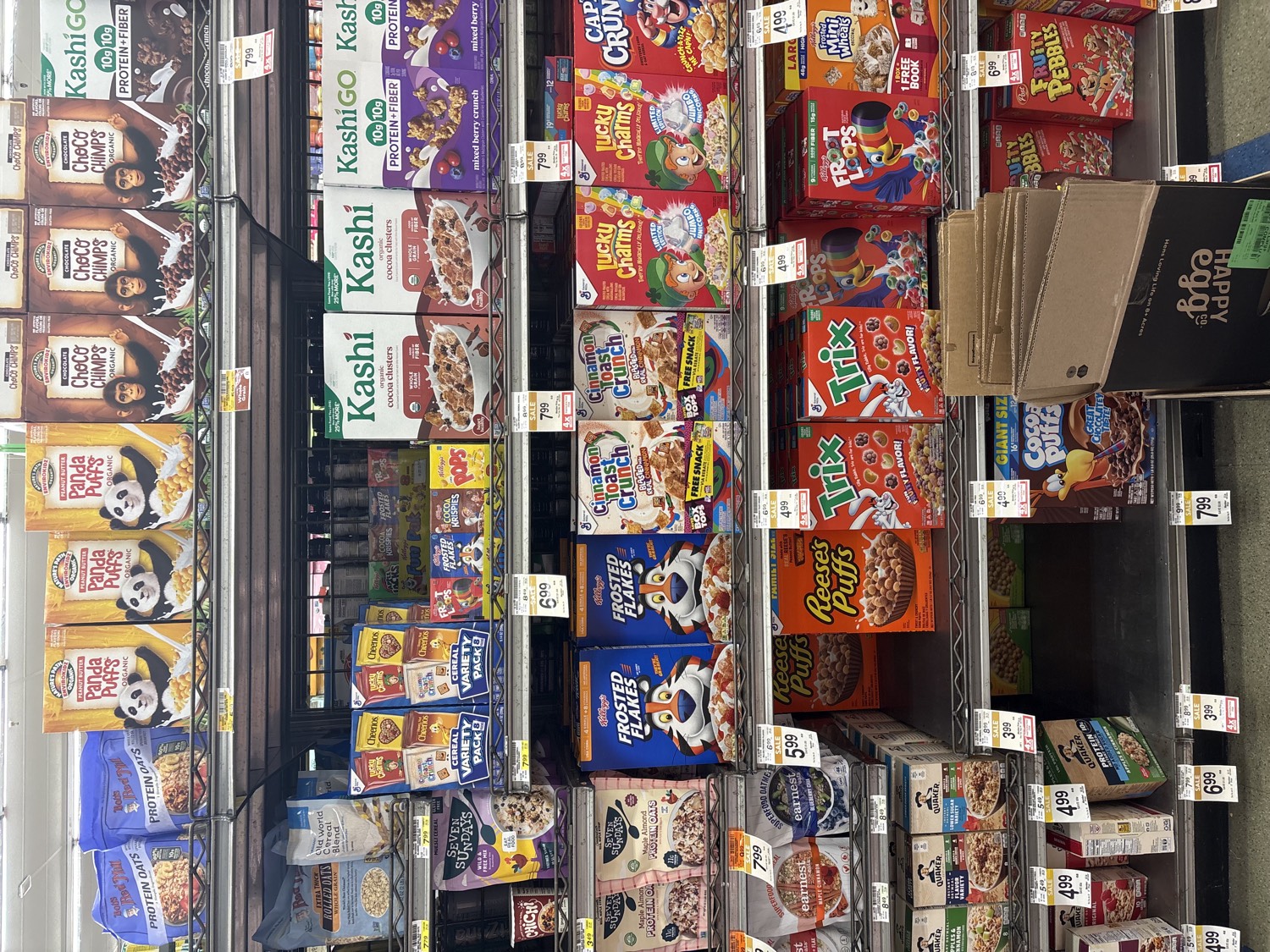 A US supermarket cereal aisle photographed at an angle, looking down the shelf: muted bran and organic health boxes such as Kashi at one end give way to brightly colored kid cereals such as Trix at the other, with yellow shelf tags throughout.
