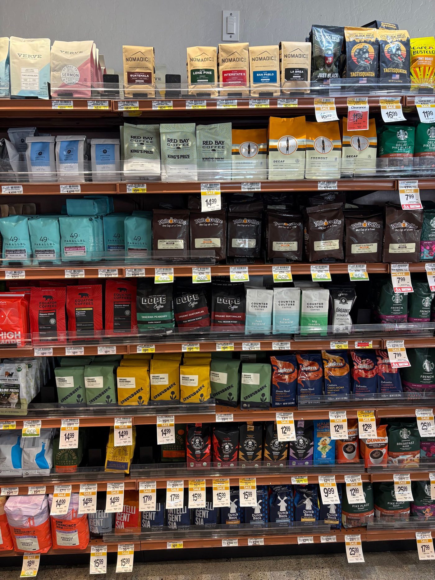 A US supermarket coffee wall photographed head-on: rows of Peet's and Starbucks bags dominating the facings, with a long tail of specialty brands below including Verve, Nomadic, Black Rifle, Red Bay, 49th Parallel, Dope, Counter Culture, Groundwork, Equator, Lavazza and Dunkin, yellow sale tags throughout.