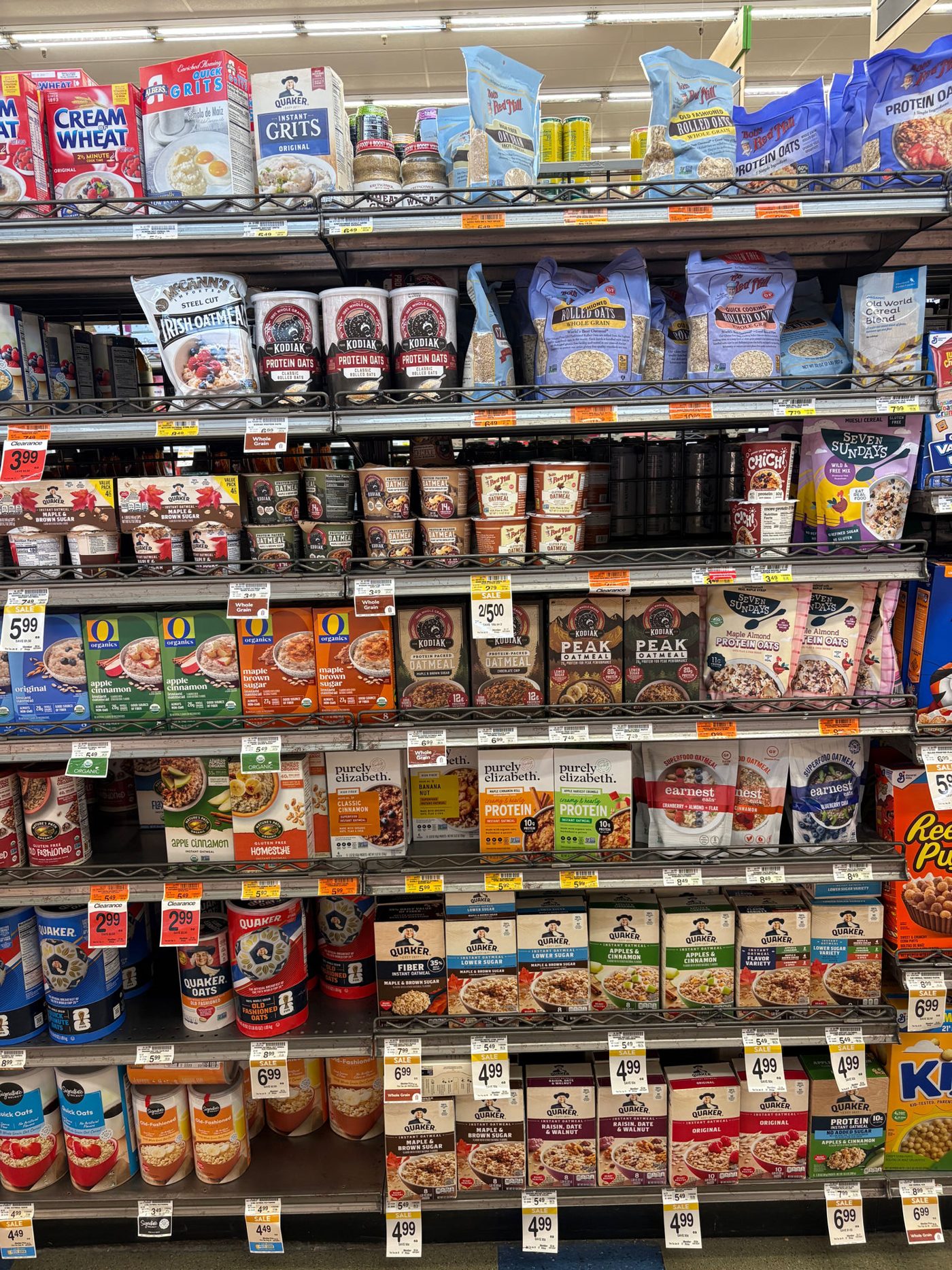 A US supermarket oatmeal wall photographed head-on: rows of Quaker canisters and instant packets, Bob's Red Mill bags, Kodiak protein oatmeal, Purely Elizabeth, Seven Sundays, McCann's steel cut, and store brands, with yellow shelf tags throughout.