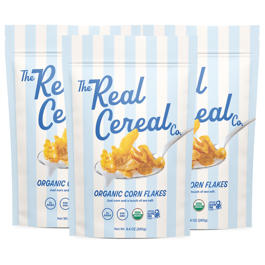 Three pale blue and white striped stand-up pouches of The Real Cereal Co. Organic Corn Flakes, a blue script logo above a photo of a spoonful of corn flakes in milk, the line "Just corn and a touch of sea salt," and badges for 0g sugar, non-GMO, USDA organic, and certified gluten-free, 8.4 oz each.
