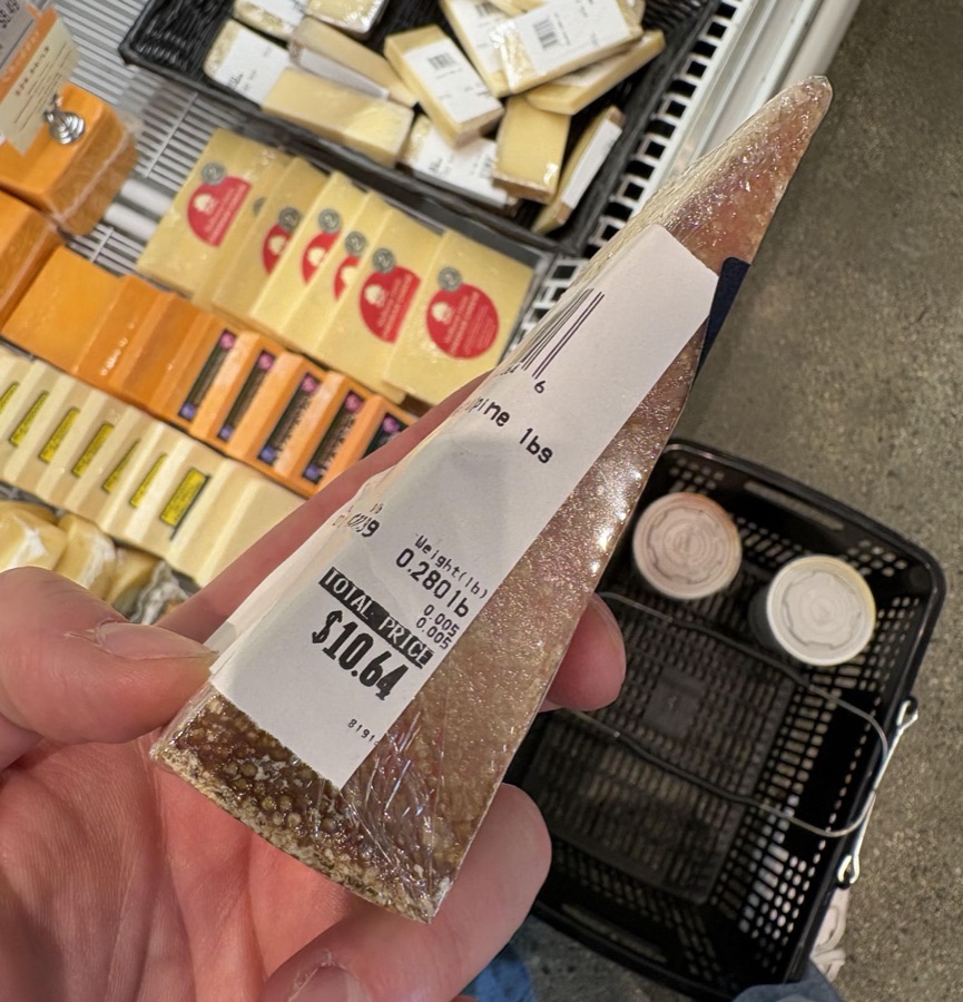A 0.28 pound wedge of alpine cheese held in front of an upscale grocery cheese case, price label reading $10.64 total