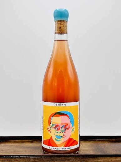 A bottle of Tinto Amorío '22 Monje skin-contact orange wine, the amber wine showing through the glass, a bold pop-art face on the label and a blue wax capsule